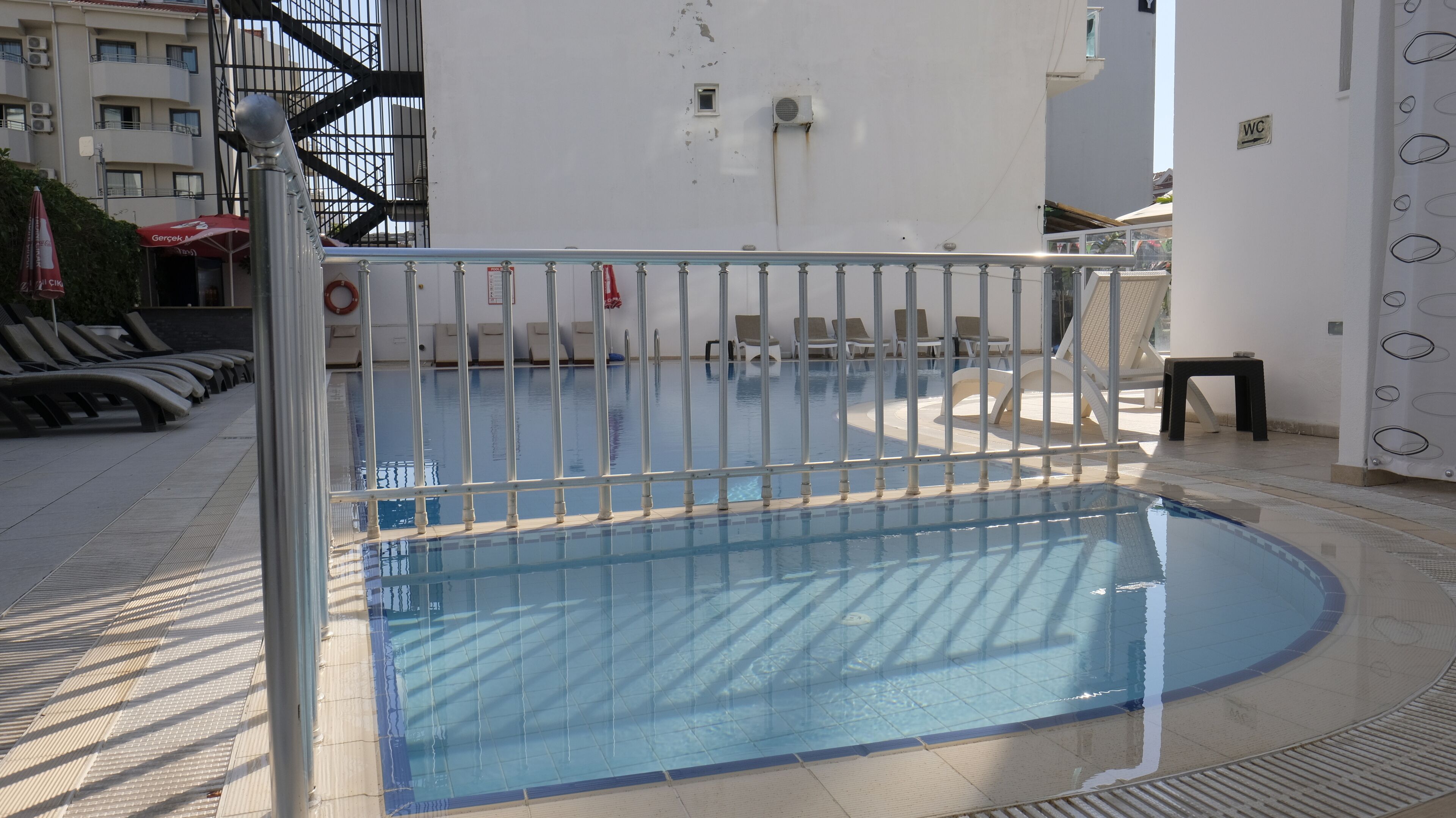 An outdoor swimming pool with a metal railing, lounge chairs, and a small table.