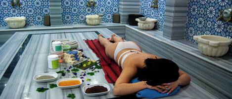 A spa bath with a person lying on a marble floor, surrounded by various bath products and a decorative towel.