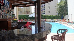 Un bar junto a la piscina con una variedad de botellas, un área de descanso con tumbonas y vista a un edificio al fondo.