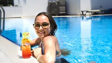 Una persona sentada junto a una piscina, con una bebida y una sombrilla.