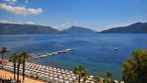 A beach with umbrellas and palm trees, boats in the water, and mountains in the background. - Golden Rock Beach Hotel - All Inclusive (Marmaris)