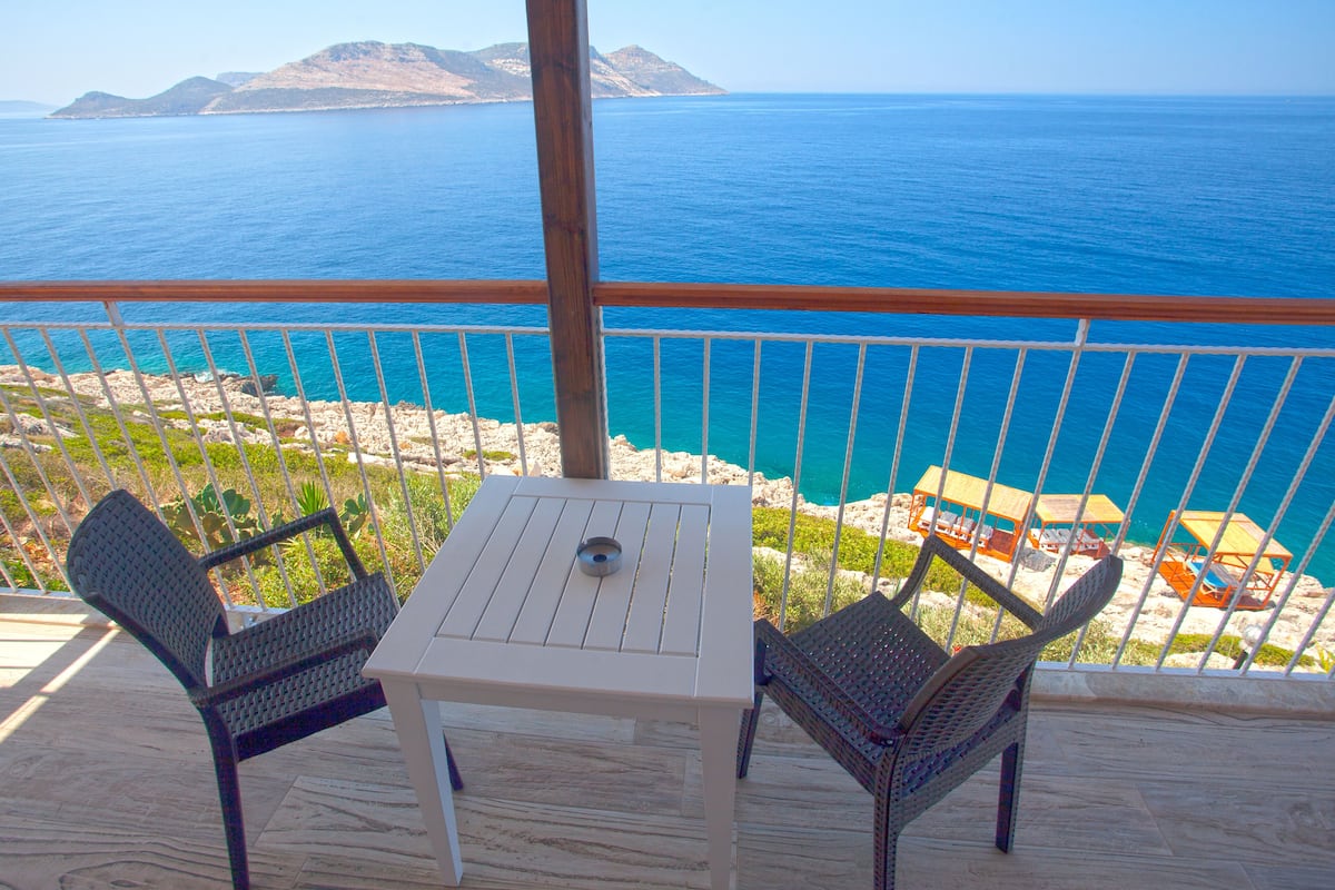 A balcony with a table and two chairs, overlooking a beach with umbrellas and a clear view of the sea and distant mountains.
