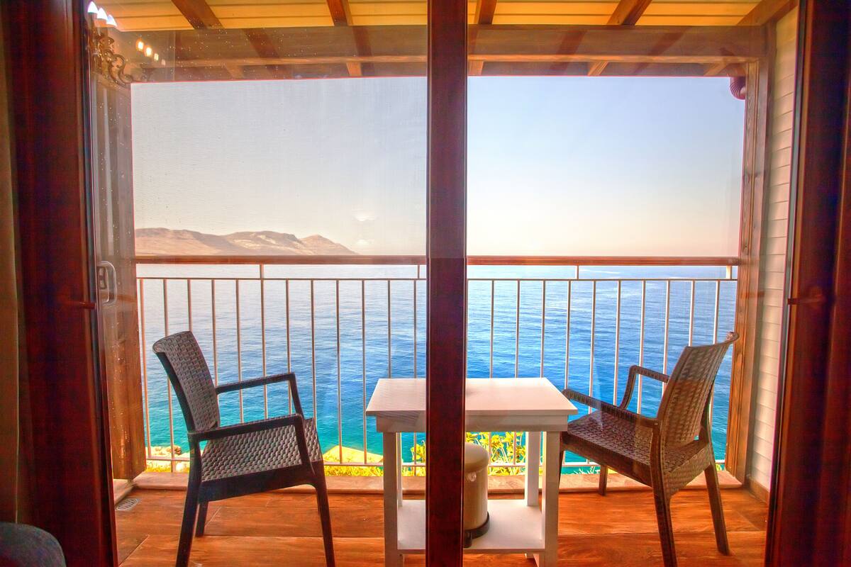A balcony with two wicker chairs and a small table, overlooking a coastal view with mountains in the distance.