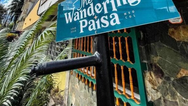 Señalización de 'The Wanderin' Paisa', con vista clara del exterior del edificio, que incluye un muro de piedra, un balcón de madera con plantas en macetas y una reja metálica.