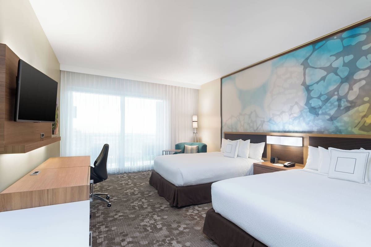 A hotel room with two beds, a desk, a chair, a TV, and a large abstract painting on the wall.
