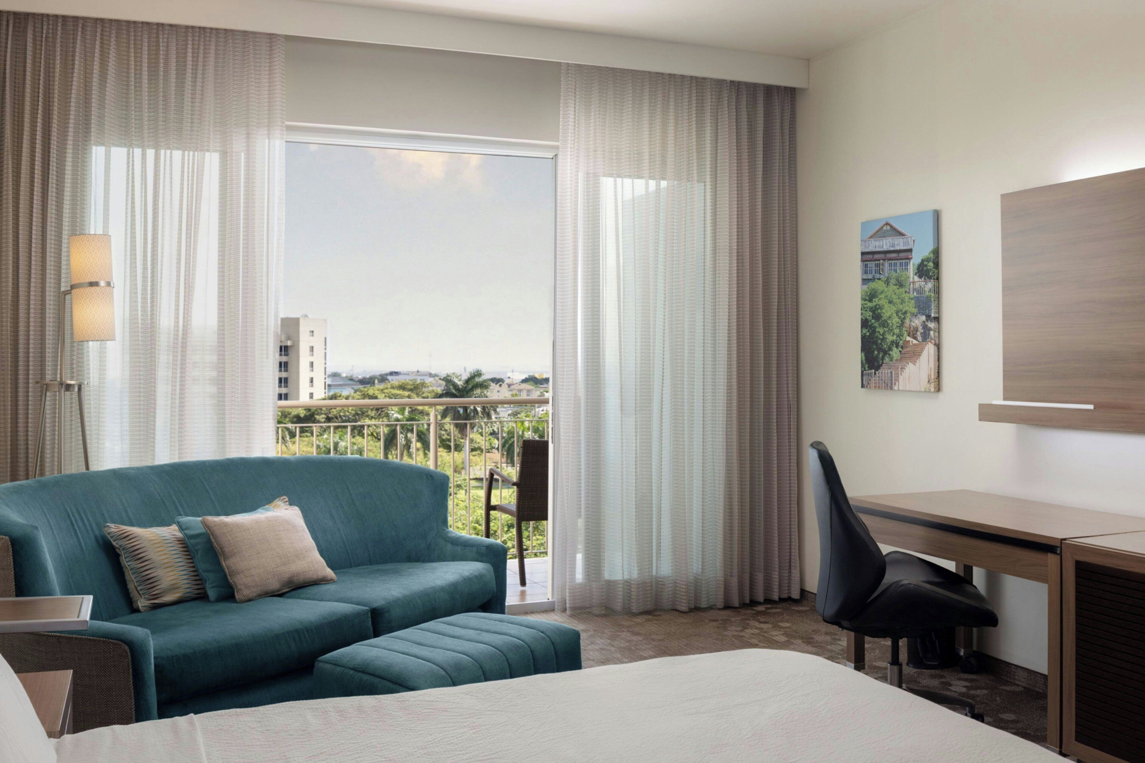 Habitación de hotel con una cama grande, un sofá, un escritorio y una silla. Tiene un balcón con vistas a edificios y árboles.