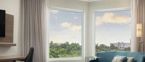 Habitación de hotel con cama, escritorio, silla, sofá y ventanas grandes que ofrecen vistas a zonas verdes y edificios.