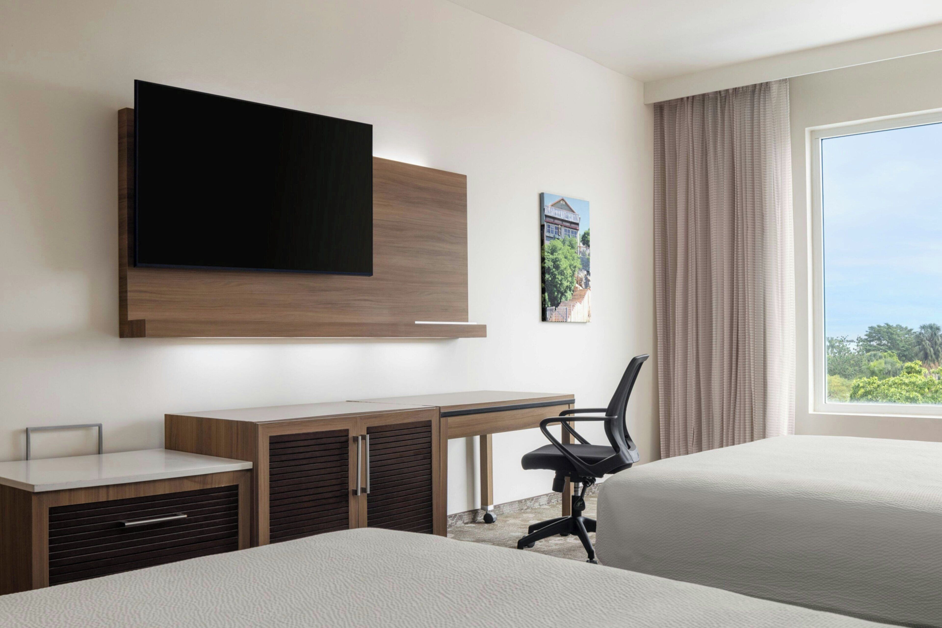 Habitación de hotel con cama, escritorio con silla, televisor montado en la pared y ventana con vistas a la vegetación.