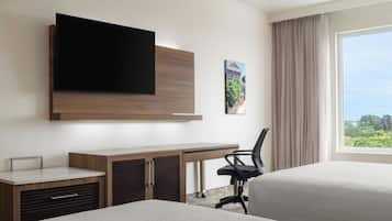 Habitación de hotel con cama, escritorio con silla, televisor montado en la pared y ventana con vistas a la vegetación.