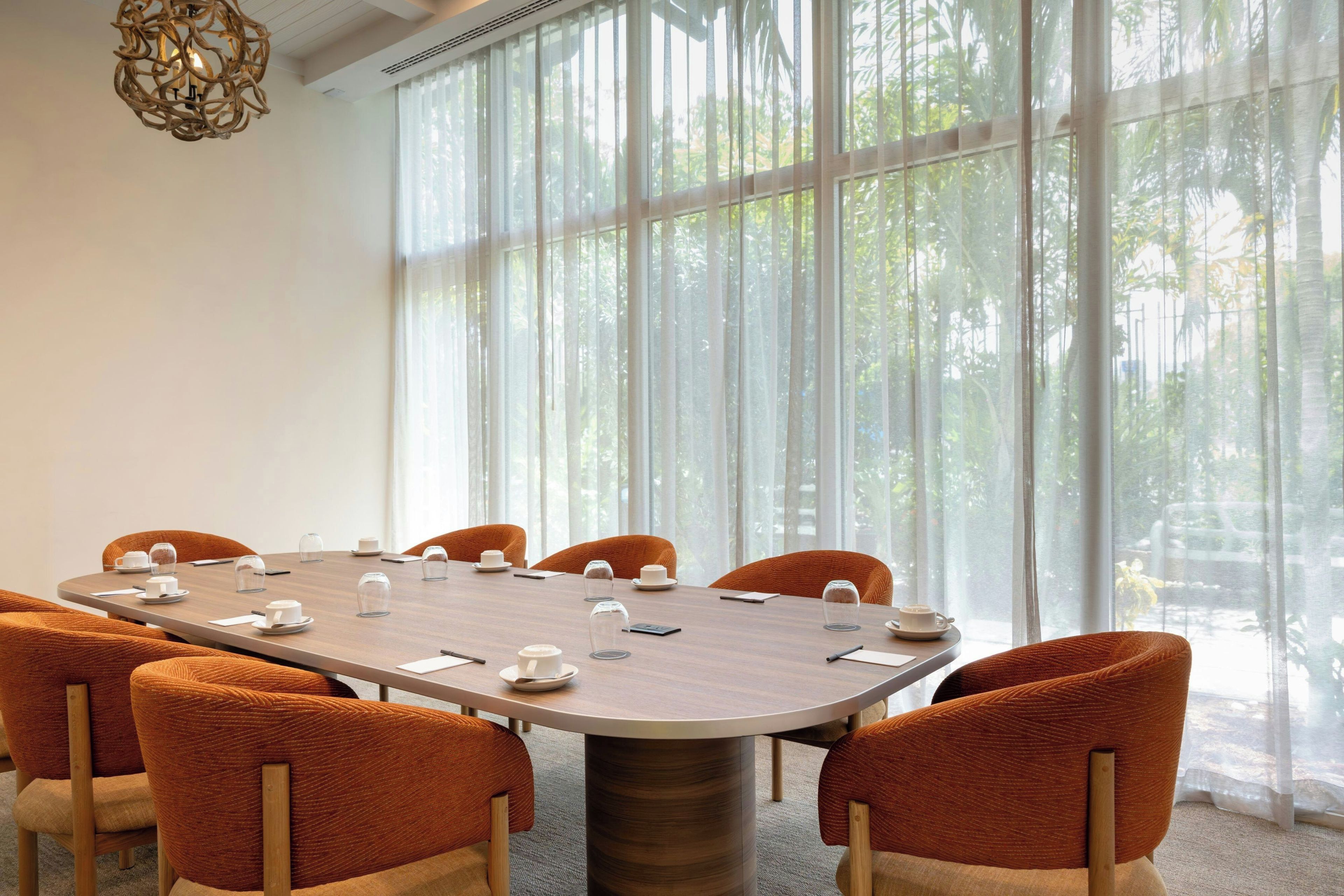 Een vergaderruimte met een ronde tafel, oranje stoelen en uitzicht op bomen door grote ramen.