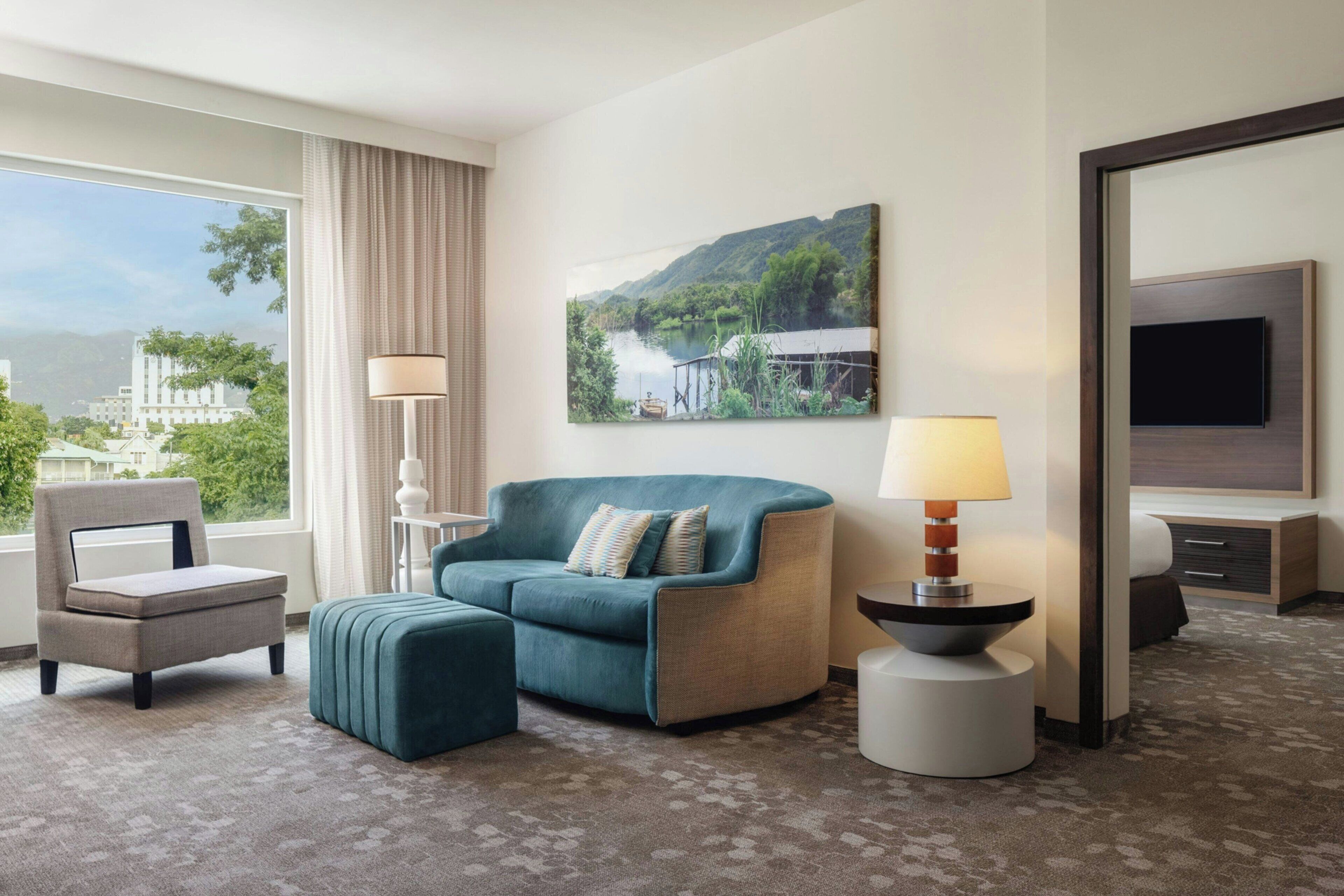 A hotel room with a sofa, armchair, and ottoman, a painting of a landscape, a lamp, and a TV.