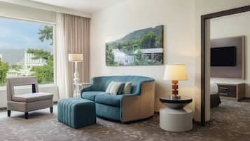 Quarto de hotel com sofá, poltrona, banqueta, pintura de paisagem, luminária e televisão.