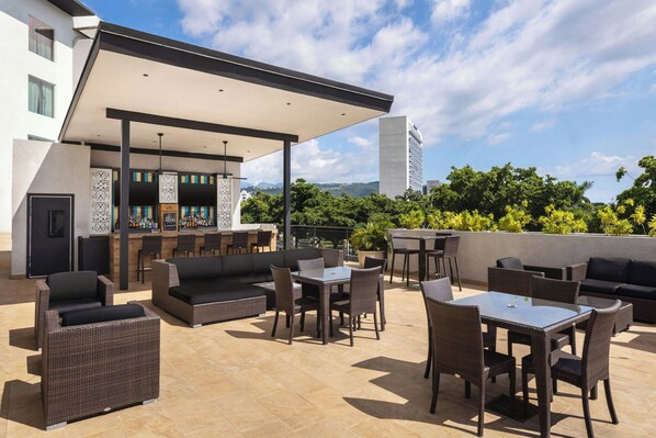 Un bar en la azotea con mesas y sofás al aire libre, con vistas a un paisaje urbano.