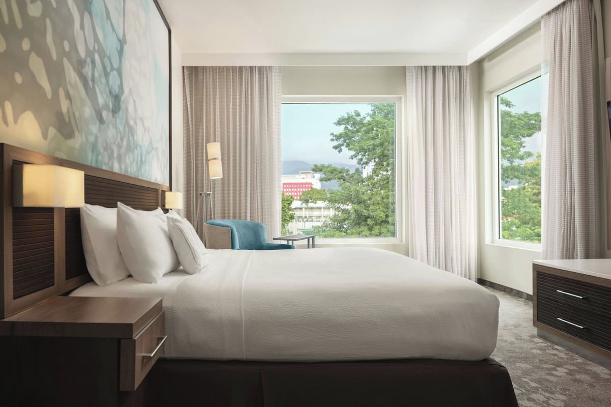 A hotel room with a large bed, a desk, a chair, and a view of the city through large windows.