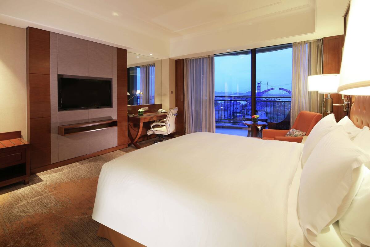Habitación de hotel con una cama grande, un escritorio con silla, un televisor en la pared y un balcón con vistas a un puente.