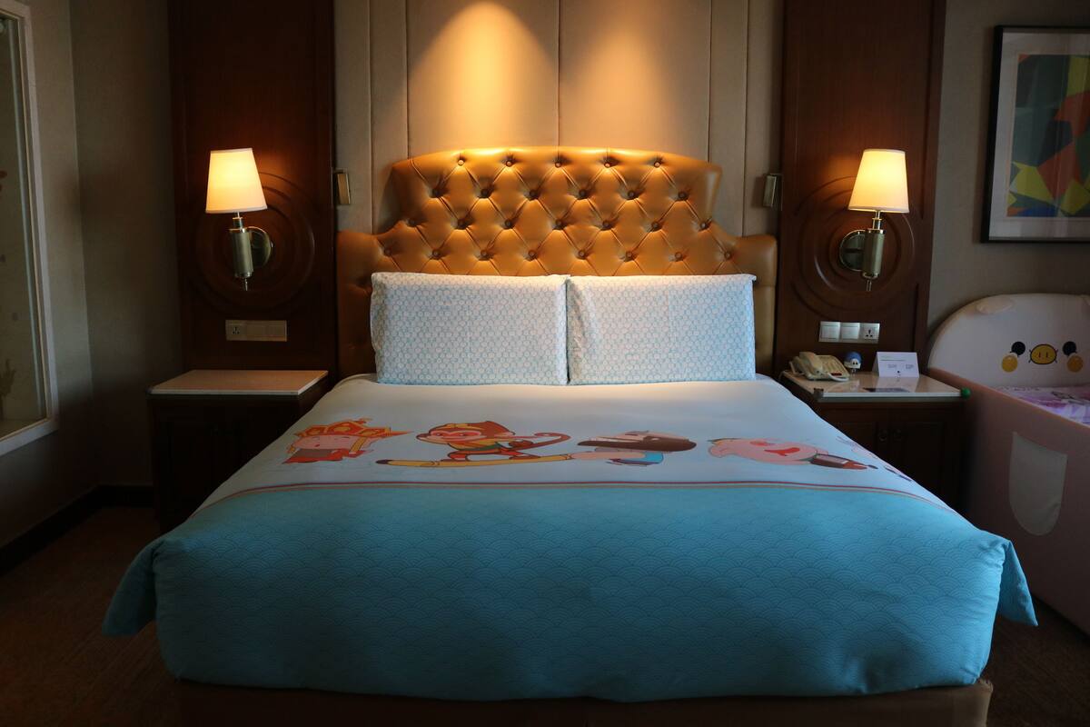 Una habitación de hotel con cabecera acolchada, dos lámparas de noche, una cama con edredón con motivos de caricaturas, una mesita de noche y un teléfono fijo en la pared.