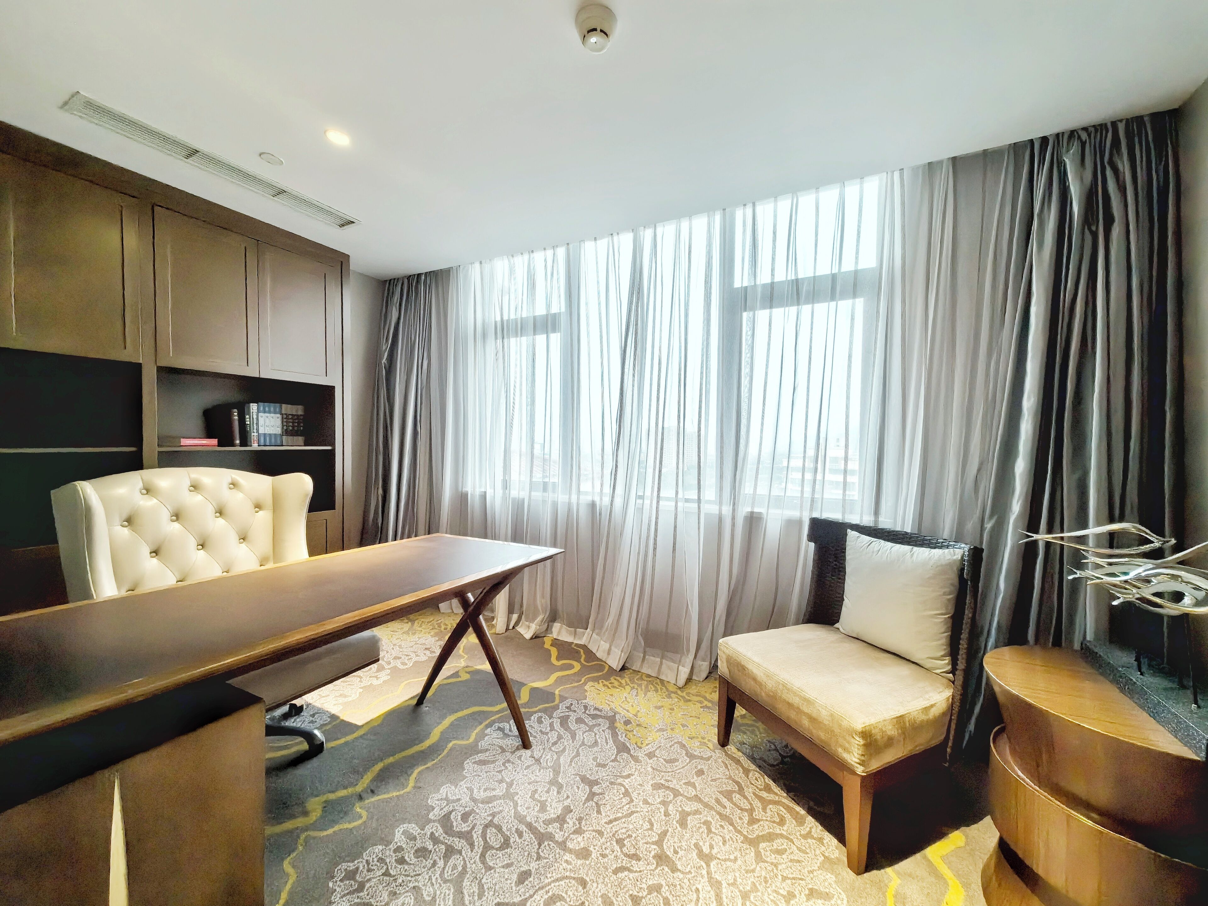 Una habitación de hotel moderna con un ventanal grande, un sillón capitonado, un escritorio de madera, un sofá y una estantería.
