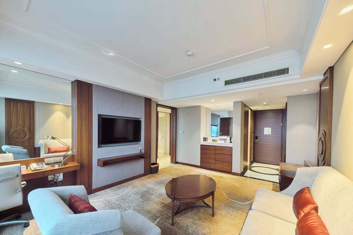 Una habitación de hotel moderna con televisión de pantalla plana montada en la pared, una zona de estar con dos sofás y una mesa de centro, un escritorio con lámpara, y un espejo grande.