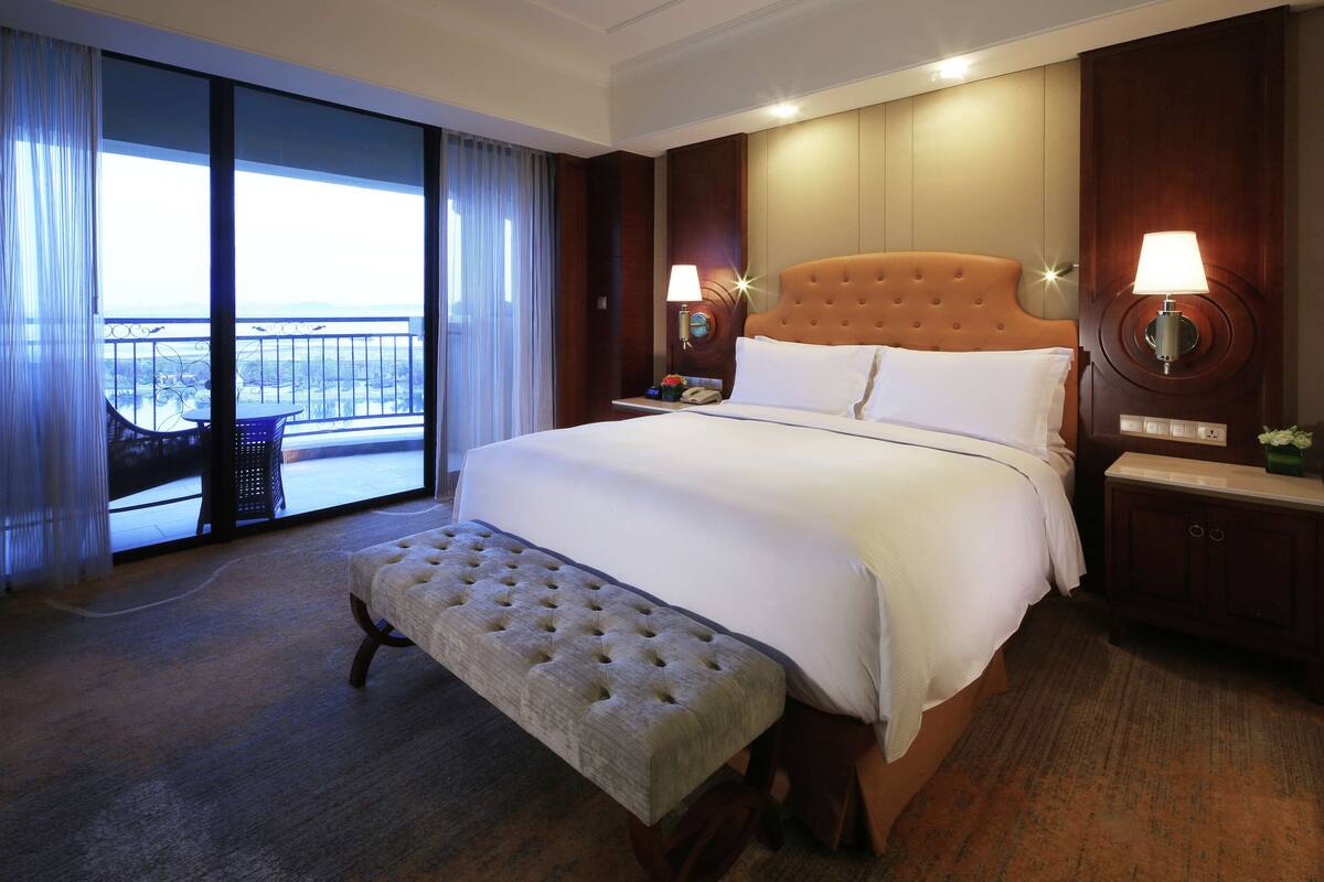 Habitación de hotel con una cama grande, un banco, una mesita de noche, una lámpara y vistas al exterior.