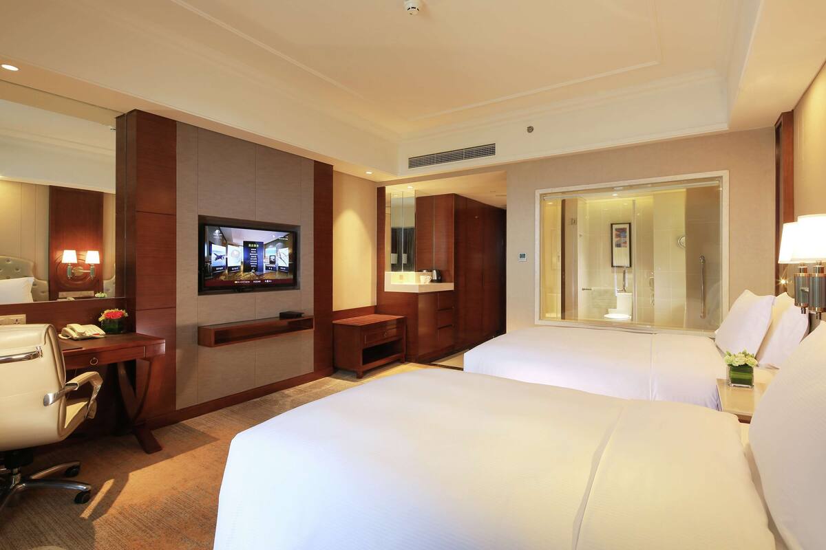 Habitación de hotel con dos camas, un escritorio con silla, televisión, baño con espejo y ducha.