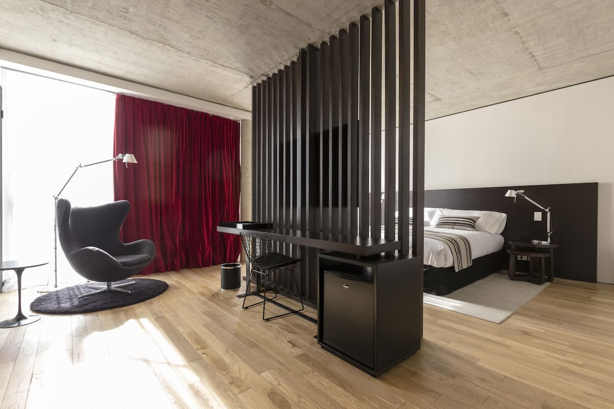 A modern hotel room with a wooden floor, a black desk, a black chair, a bed with white bedding, and a red curtain.
