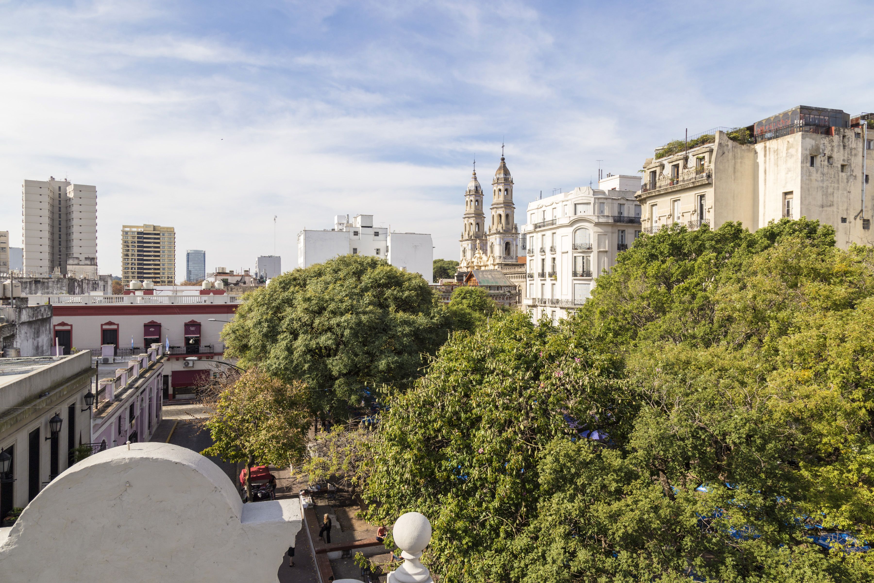 A cityscape with historic buildings, a prominent church with two towers, and lush green trees in the foreground.