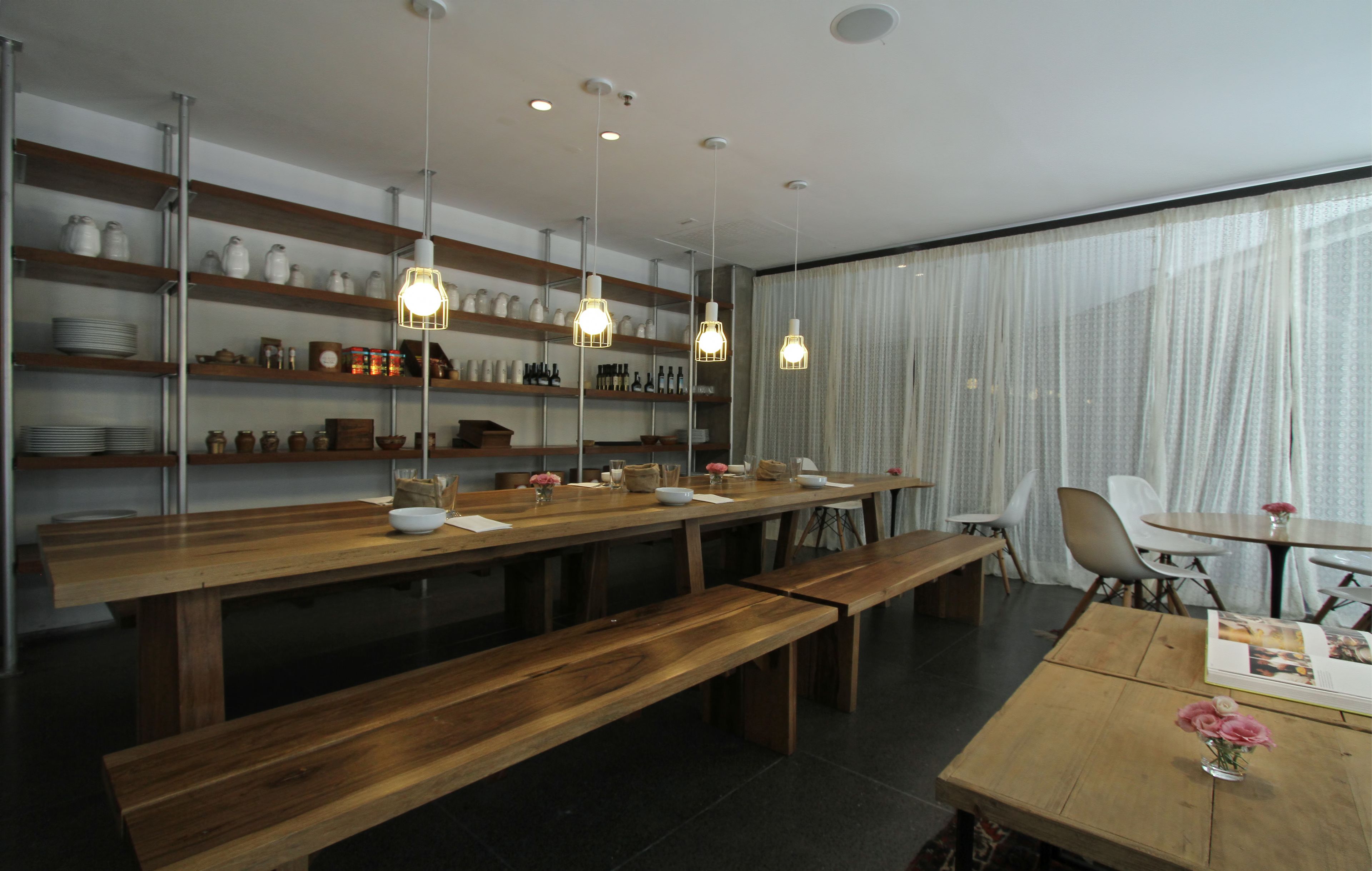 A modern dining area with wooden tables and benches, shelves with kitchenware, and pendant lighting.