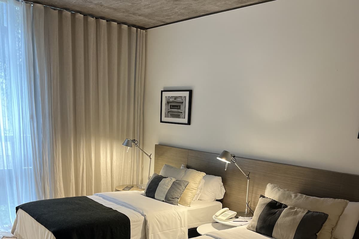 A hotel room with two beds, a bedside table with a phone, and a window with curtains.