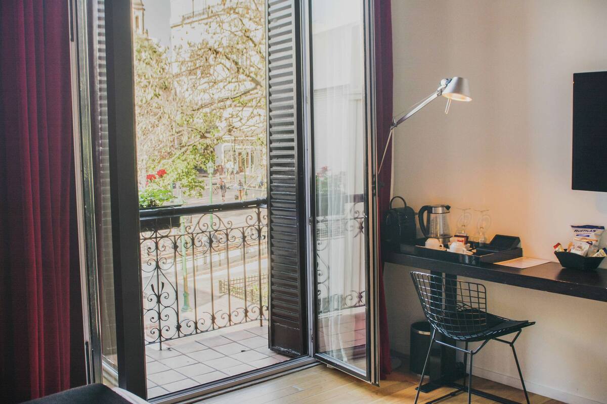 A room with a sliding glass door leading to a balcony, a desk with a chair, a lamp, and a coffee maker.