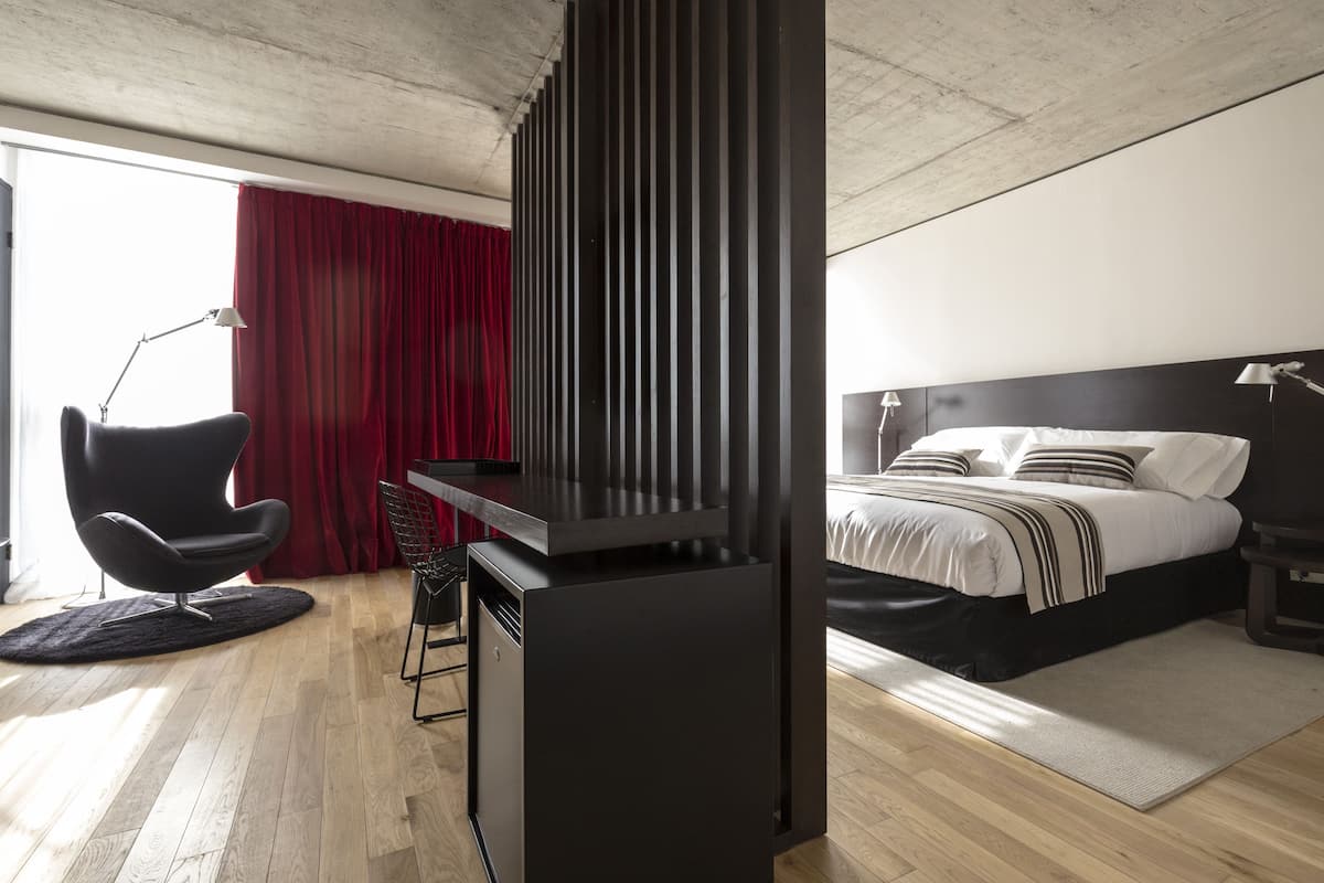 A modern hotel room with a wooden floor, a black desk with a chair, a bed with white bedding, and a red curtain.