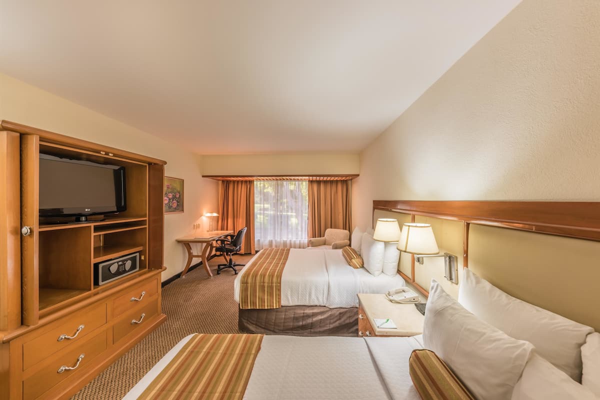 A hotel room with a TV, desk, two beds, lamps, and a window with curtains.