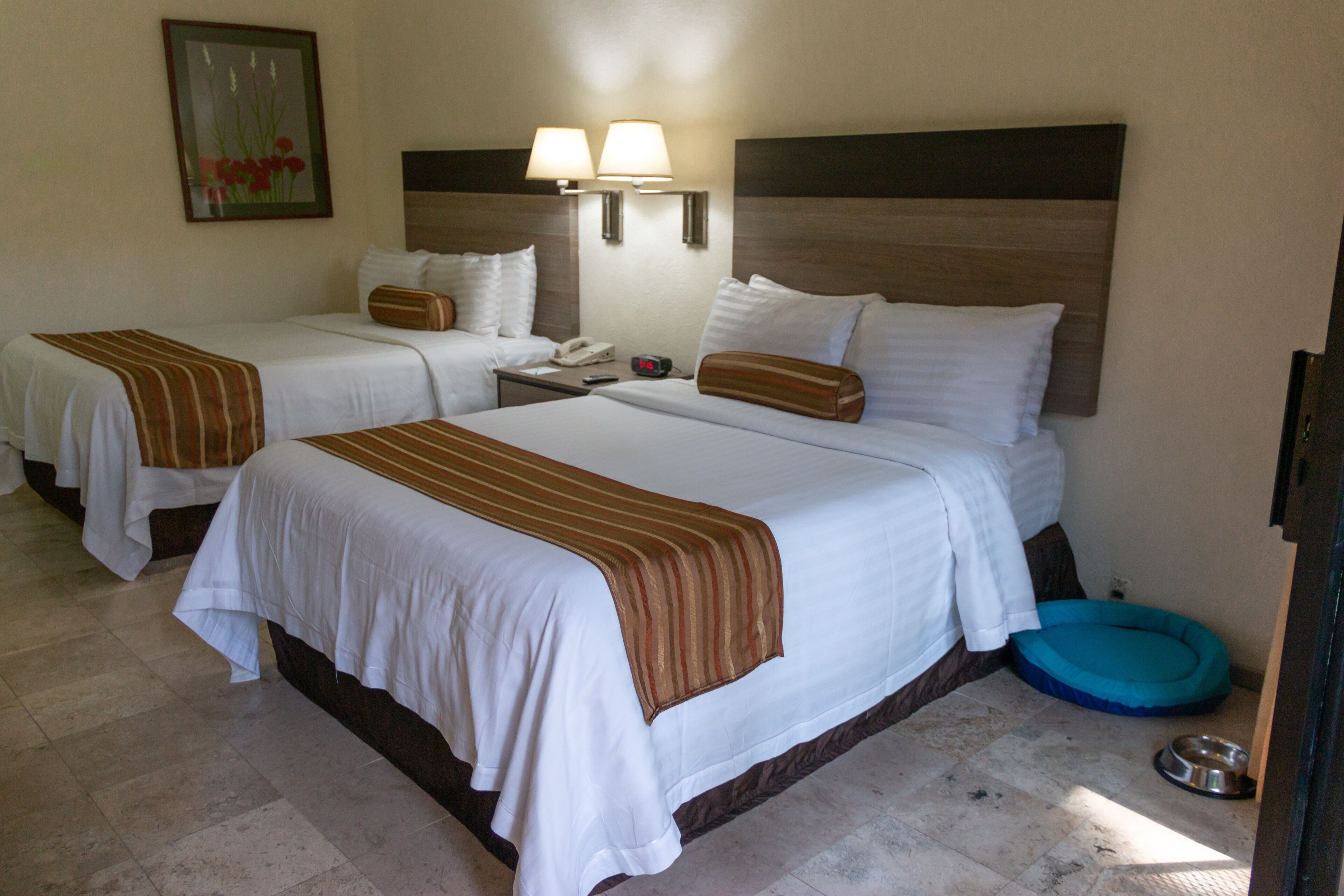 A hotel room with two beds, a nightstand, a lamp, a painting, and a pet bed.