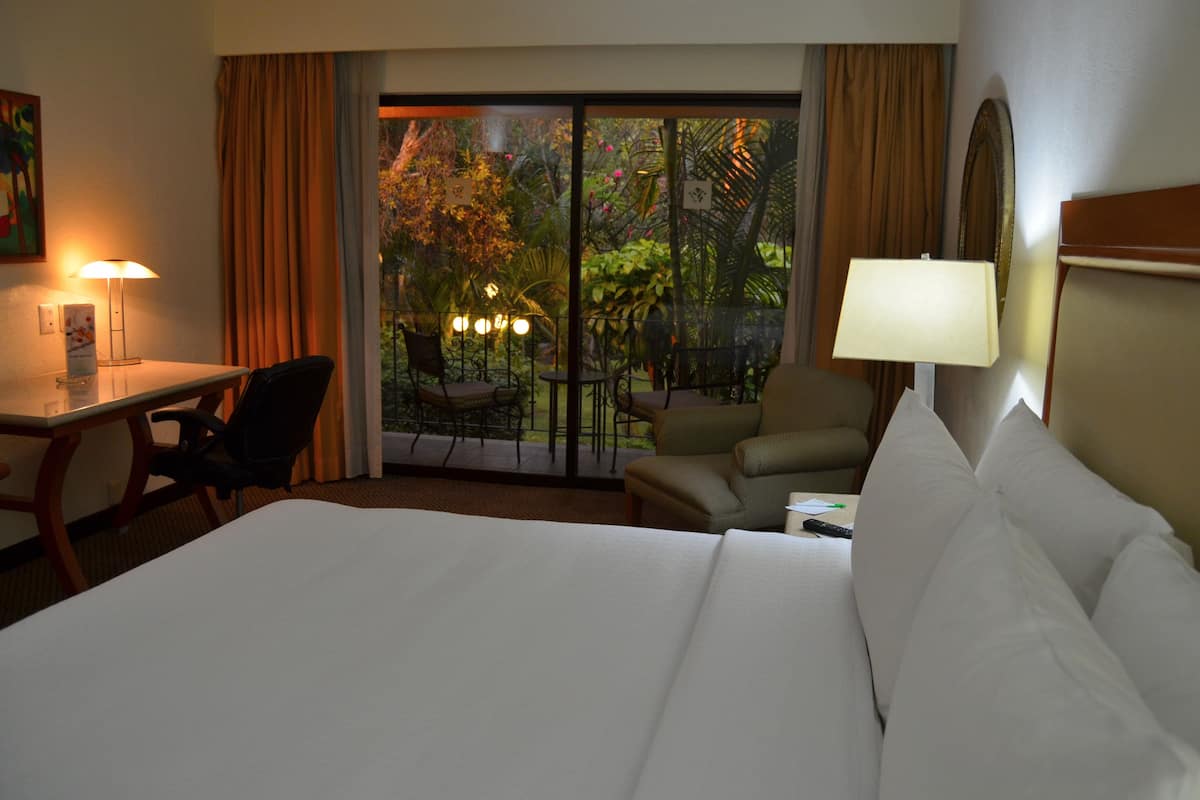 A hotel room with a large bed, a desk, and a chair. There is a balcony with a view of trees and a garden.