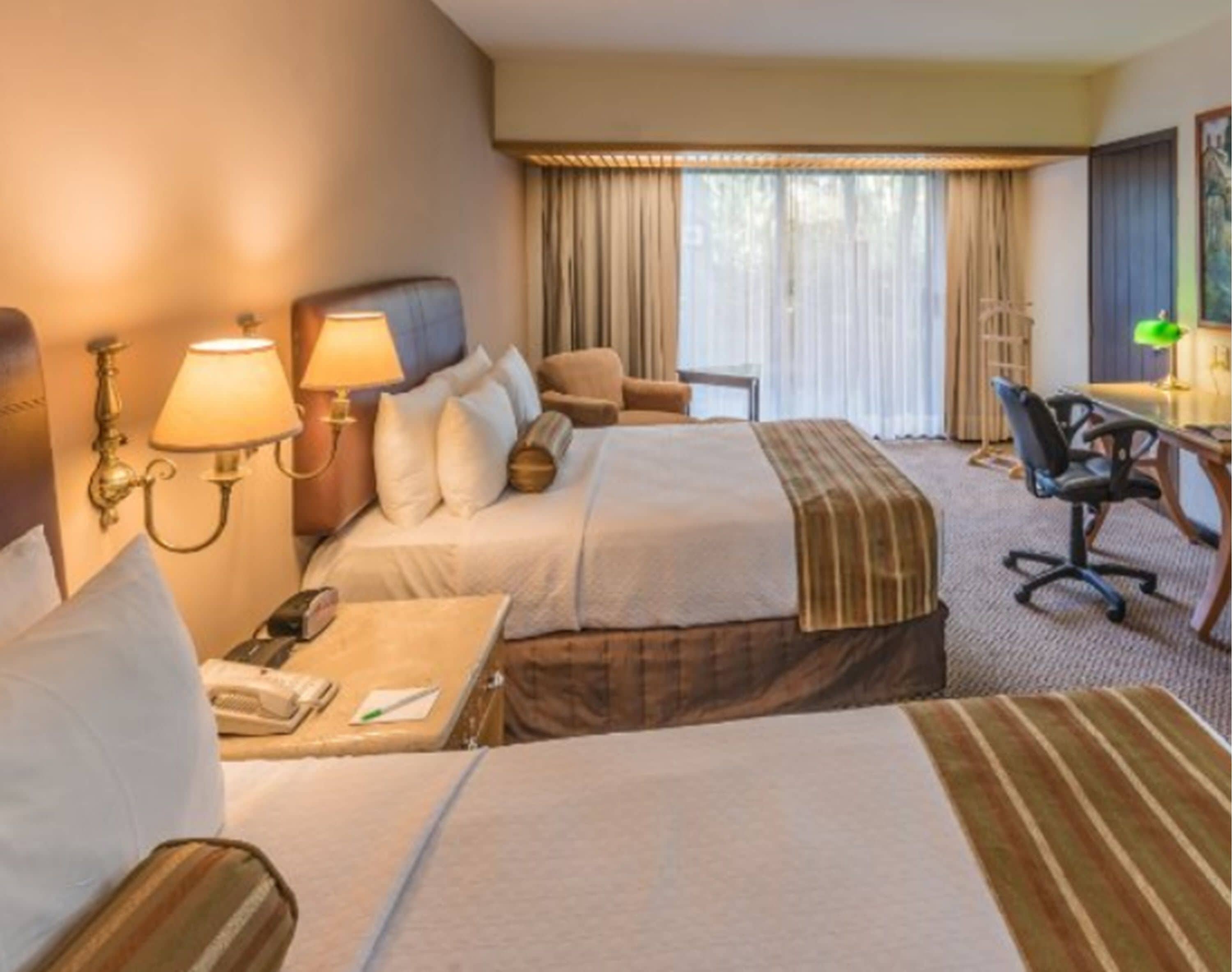 A hotel room with two beds, a desk, a chair, and a lamp.