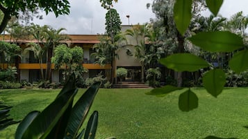 Un edificio con una entrada central, rodeado de un césped cuidado y diversas plantas tropicales.