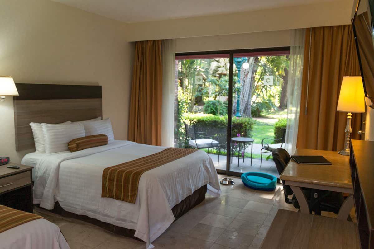 A hotel room with a large bed, a desk, a chair, and a view of a garden through a sliding glass door.
