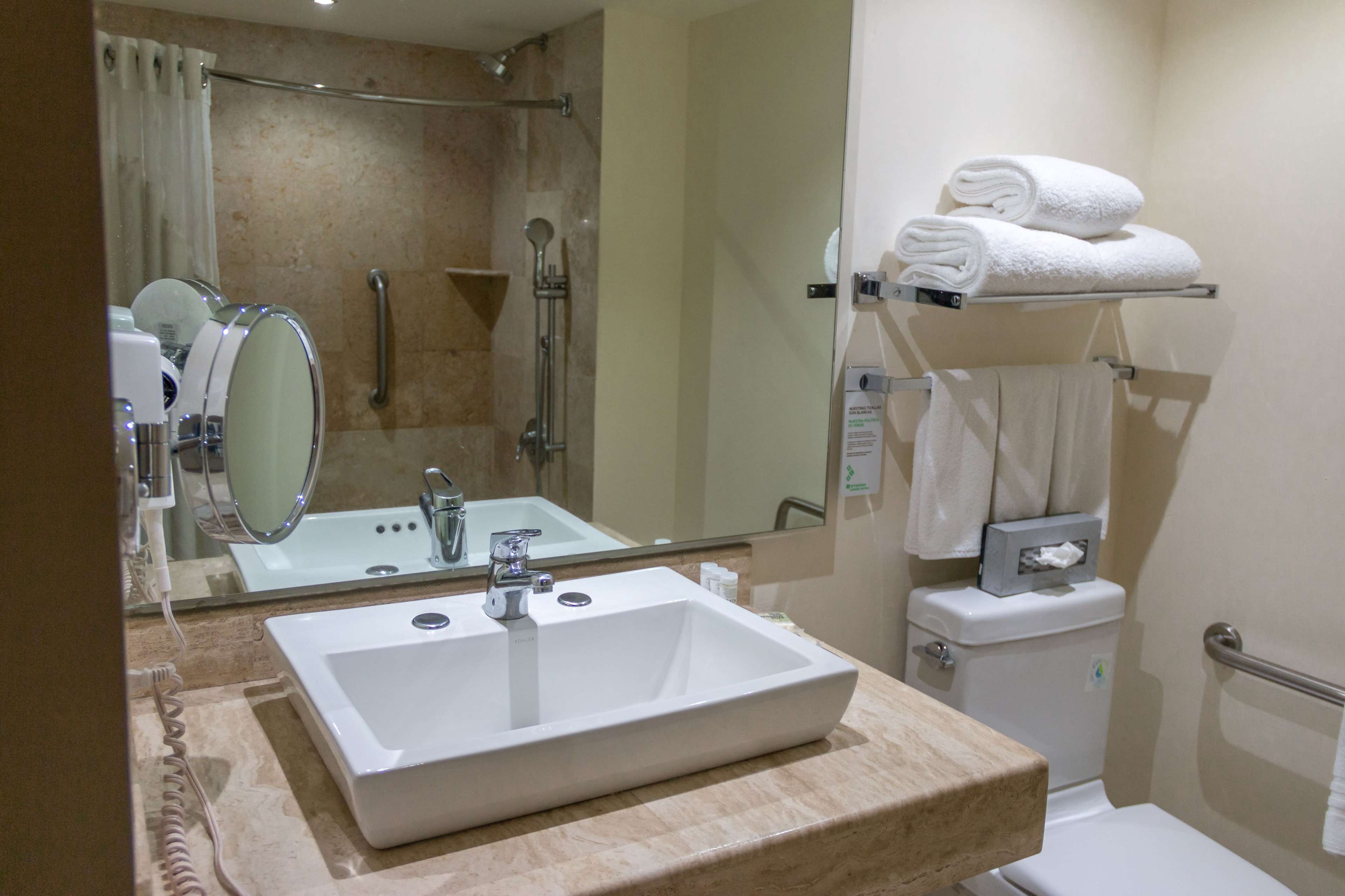A bathroom with a large mirror, a sink, a shower, and a toilet. There are towels on a rack and a handrail near the toilet.