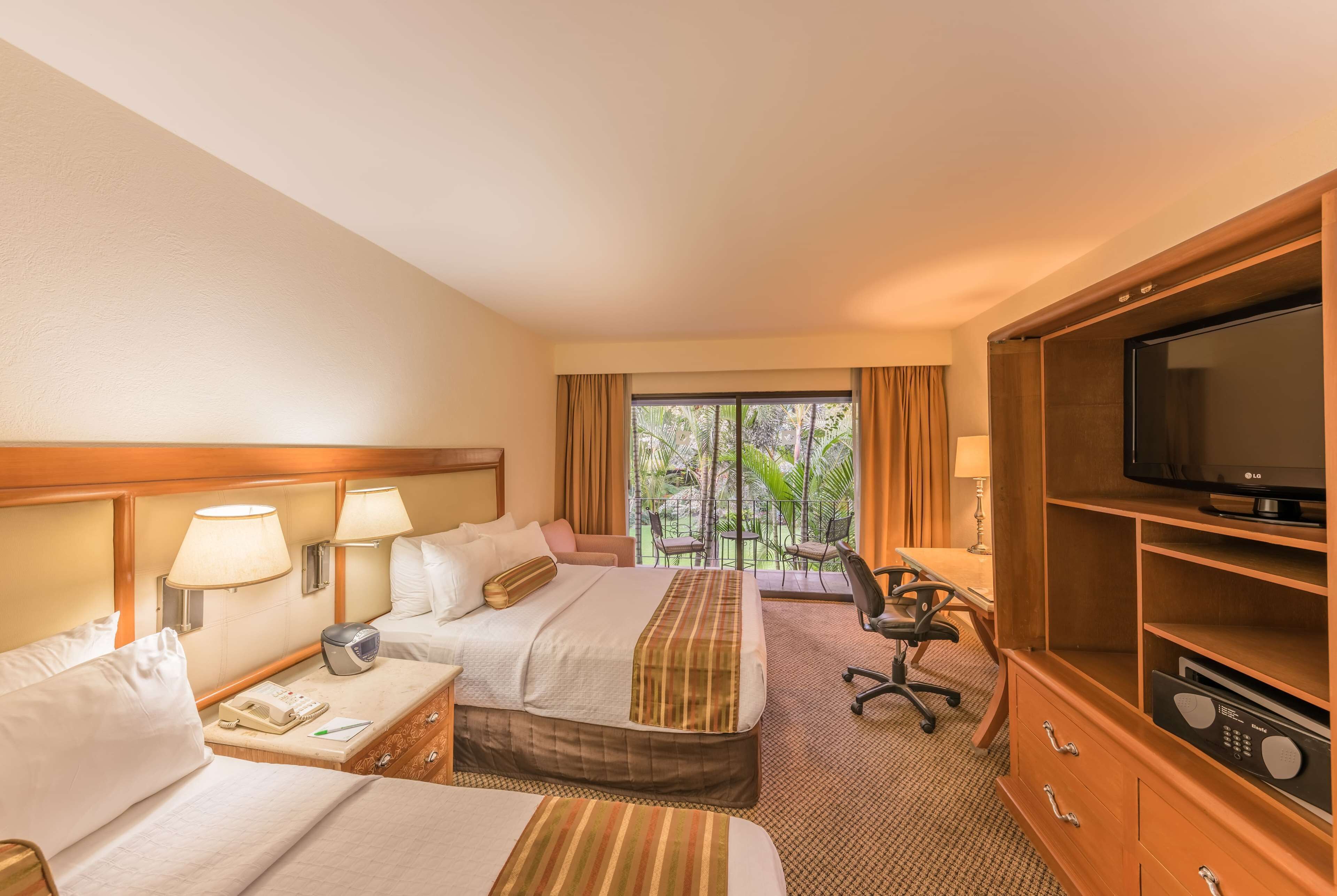 A hotel room with two beds, a desk, a TV, and a balcony with a view of greenery.