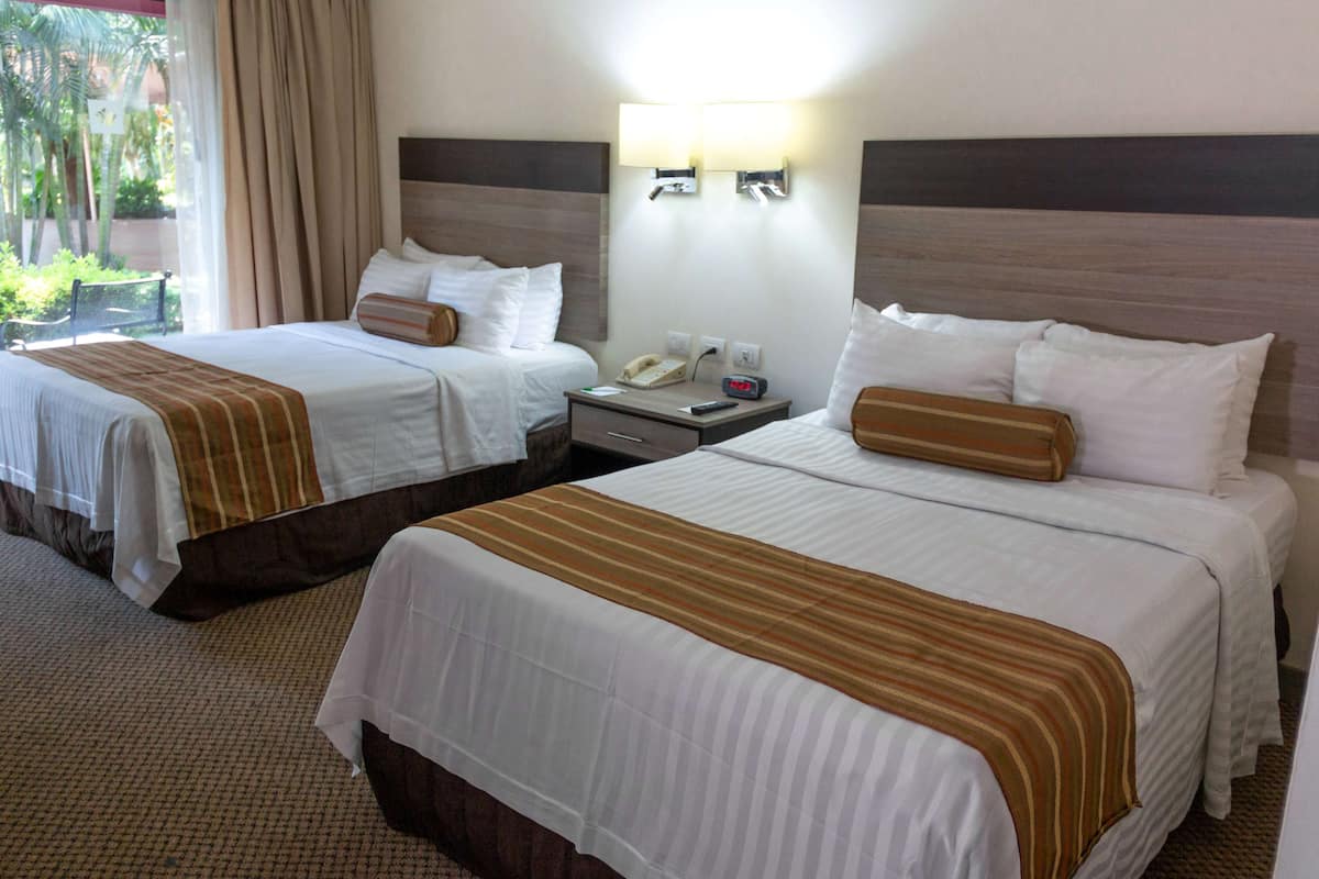 A hotel room with two beds, each with a striped bedspread, a nightstand with a phone and a clock, and a wall-mounted light fixture.