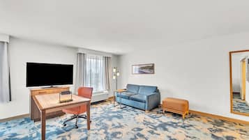 A hotel room with a wooden desk, a blue sofa, a small orange ottoman, a flat-screen TV, and a large mirror.