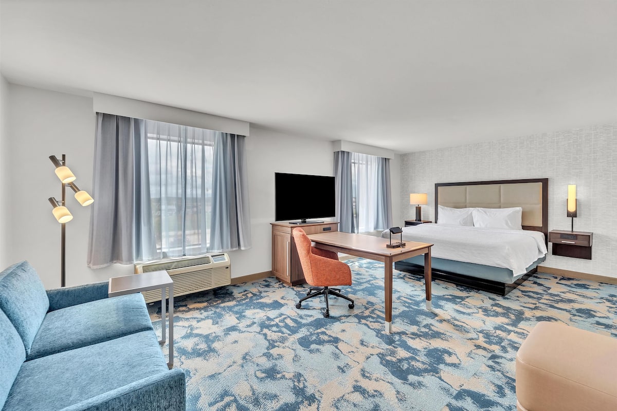 A hotel room with a bed, desk, chair, TV, and a large window with curtains.