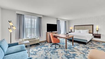 Una habitación de hotel con cama, escritorio, silla, televisor y un amplio ventanal con cortinas.