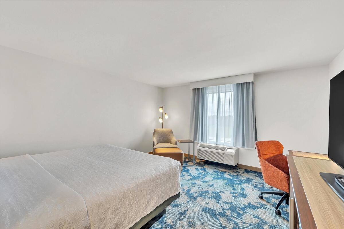 A hotel room with a bed, a desk, an orange chair, a small table, and a window with curtains.