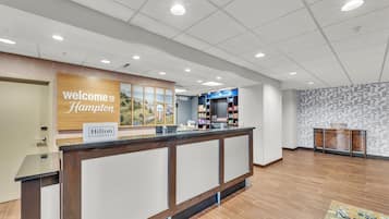 Un área de recepción de hotel con un mostrador de madera, un letrero que dice 'Welcome to Hampton', un letrero que dice 'Hilton Honors' y una pared con diseño.