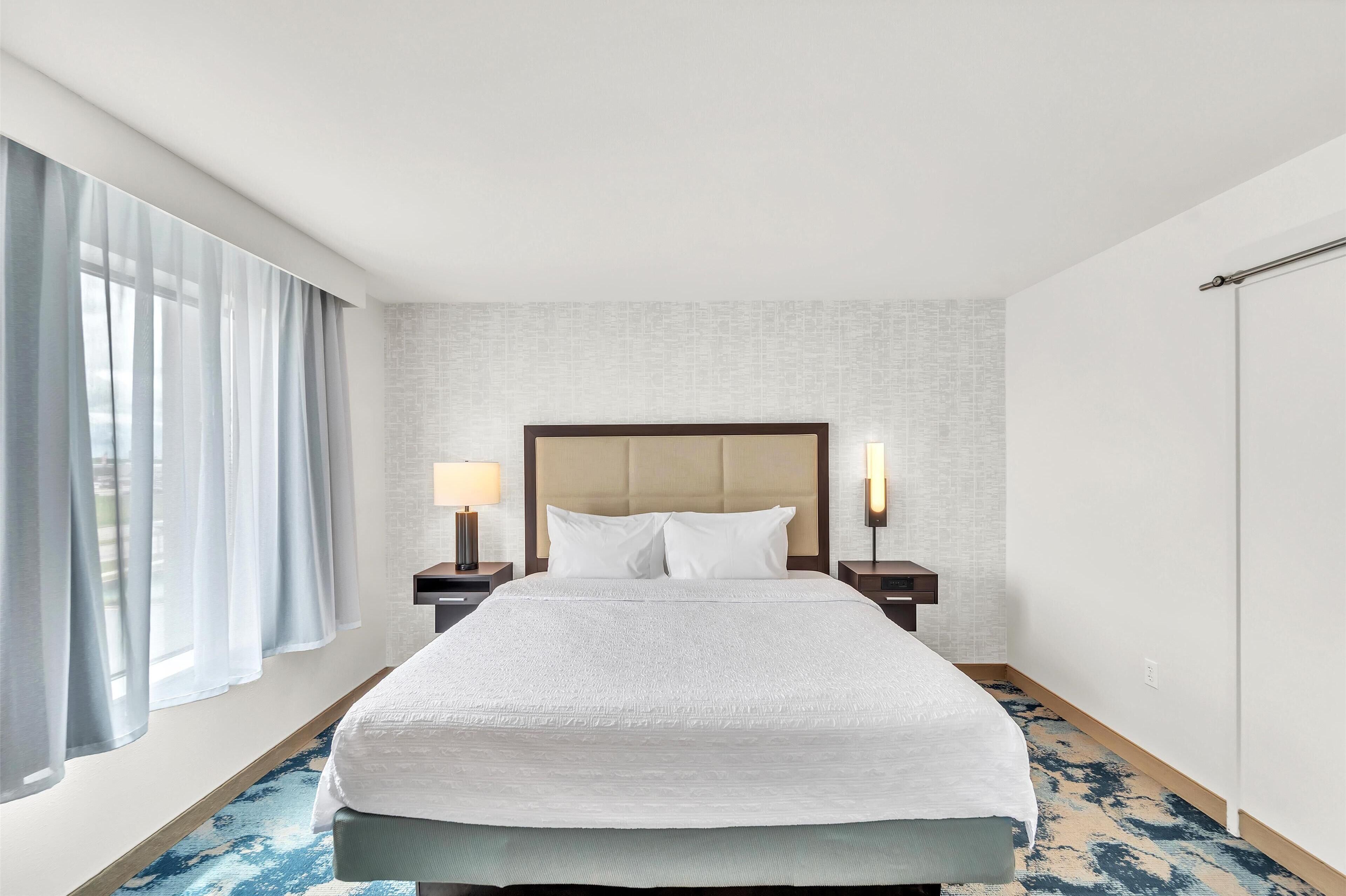 Ein Hotelzimmer mit einem großen Bett, zwei Nachttischen mit Lampen, einem Fenster mit Vorhängen und einem gemusterten Teppich.