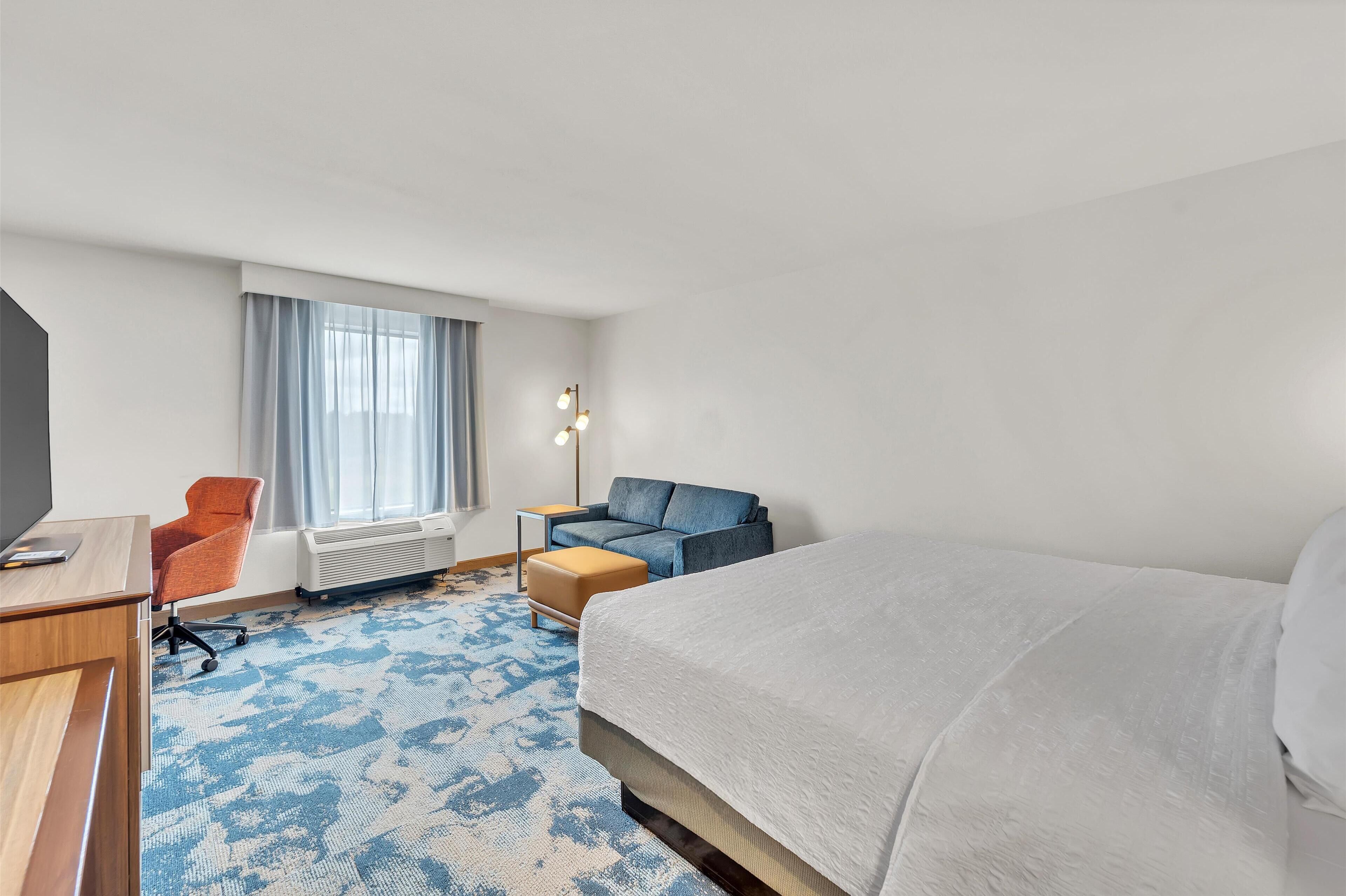 A hotel room with a large bed, a desk, an orange chair, a blue sofa, and a window with curtains.