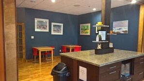 A kitchen area with a granite countertop, wooden cabinets, and a trash bin. There are two red chairs and a wooden table. The walls are painted blue, and there are framed artworks on the wall. - Marble Inn Resort (Steady Brook)