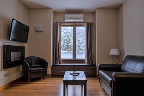 A living room with a sofa, a chair, a coffee table, a TV, and a window with a view of snow-covered trees. - Marble Inn Resort (Steady Brook)