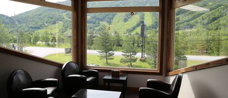 A room with large windows offering a view of a green valley and a winding road. There are several black leather chairs and a small table.