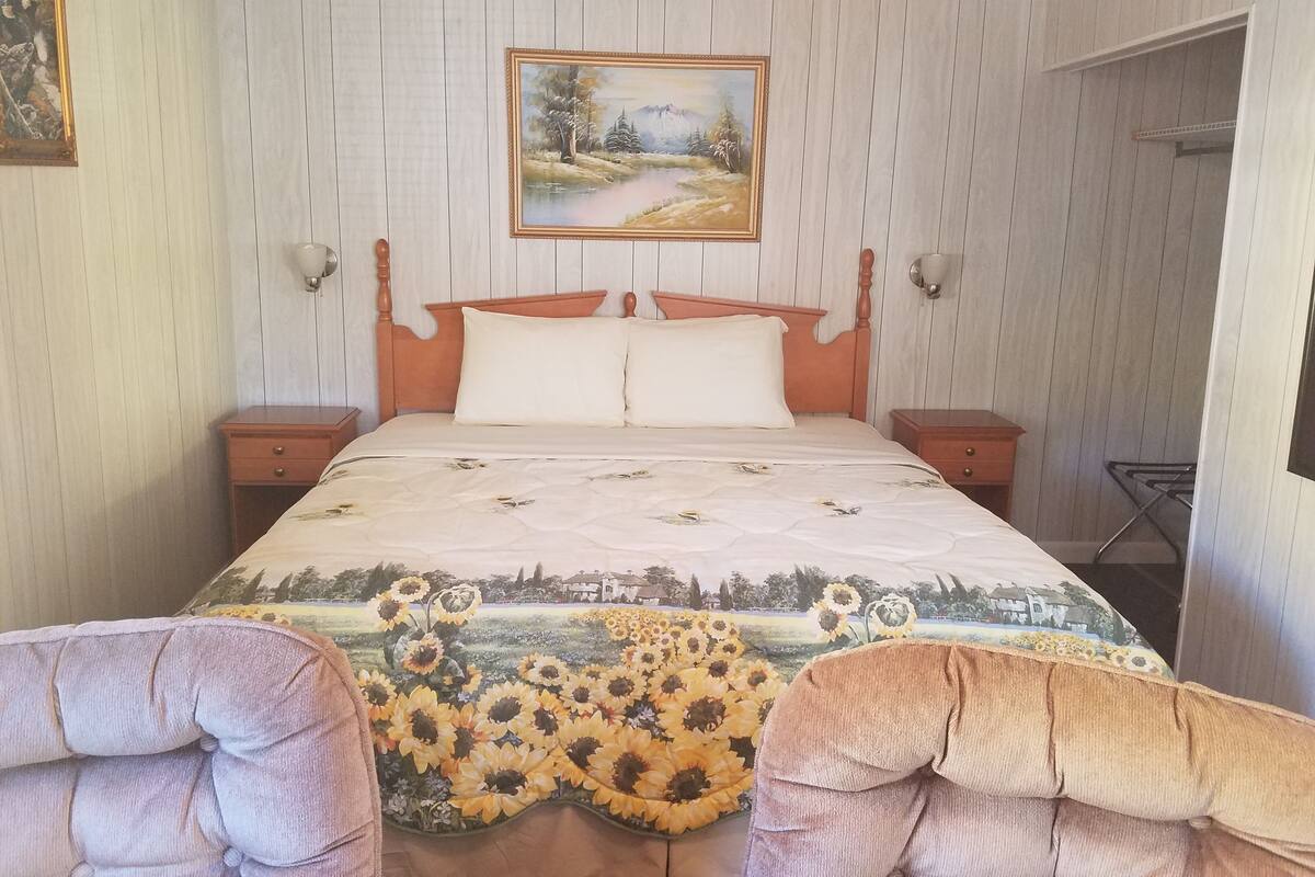 A bedroom with a wooden bed, two bedside tables, a painting above the bed, and a sunflowers-themed bedspread.